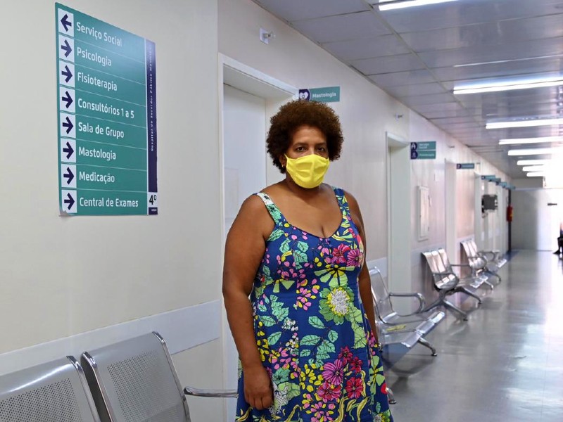 Foto da professora Lídia. Ela está parada no meio do corredor do hospital, olhando para a foto, e usa uma máscara amarela e vestido florido